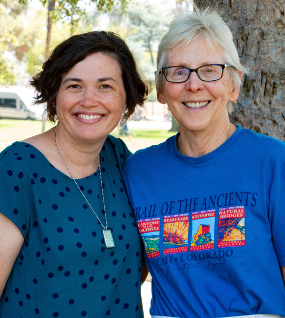 Two women stand arm in arm smiling broadly at the camera