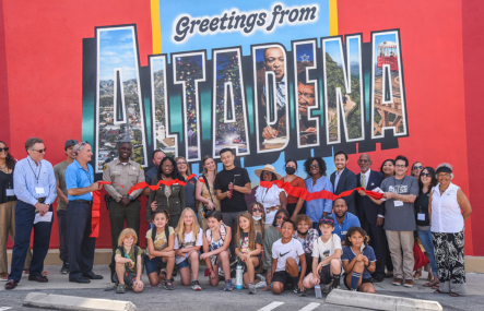 A crowd of more than a dozen people stand in front of a vibrant red wall with a mural that says Greetings From Altadena