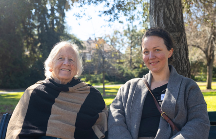 Two women sit together outdoors with trees and grass visible behind
