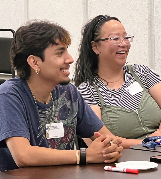 A man and woman sit side by side at a table with papers in front of them, they are laughing together and looking to the side