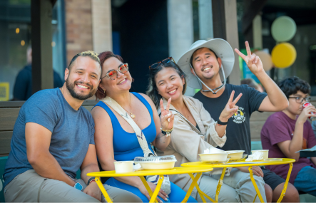 Four adults sit closely together outdoors with bright yellow tables in front of them. Two of the people hold up peace signs with their fingers