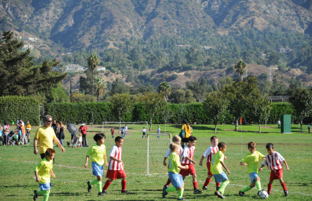 A long shot shows several children practicing soccer in a park with large mountains behind them