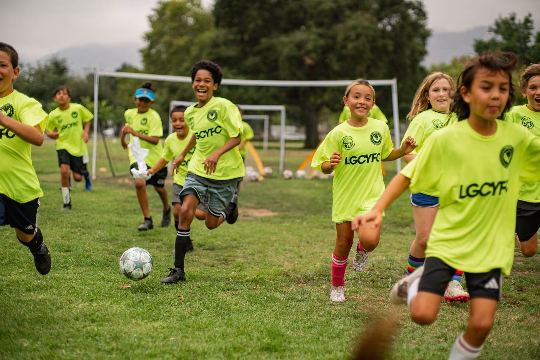 A group of elementary school-aged children all wearing the same bright green soccer jerseys, run on a soccer field toward the camera, laughing and kicking the ball
