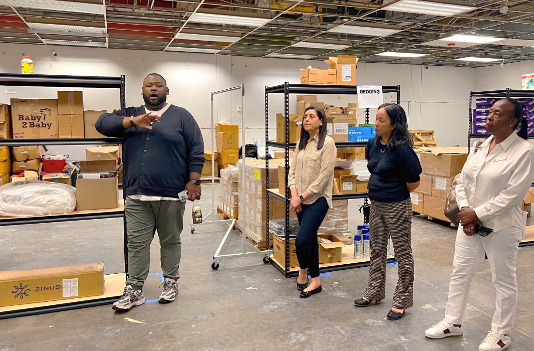 A man stands inside a supply warehouse, speaking and gesturing; Three adult females stand on the right of the photo listening to him.