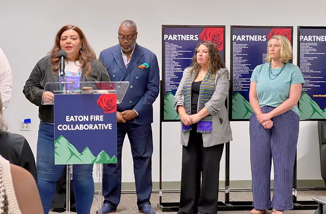 A woman stands at a podium that says Eaton Fire Collaborative. Behind her are three adults standing and listening, and three signs that say Partners
