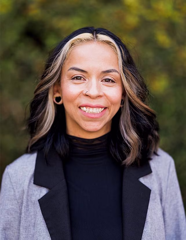 A woman is smiling in a black shirt and blazer with a green leaf background.