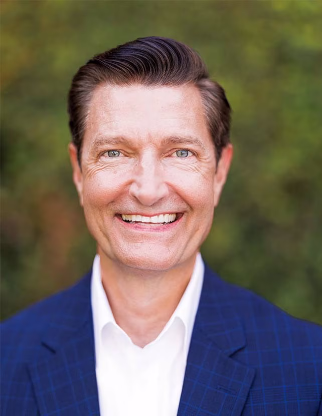 A man is smiling in a blue blazer with a green leaf background. He is wearing a white shirt.