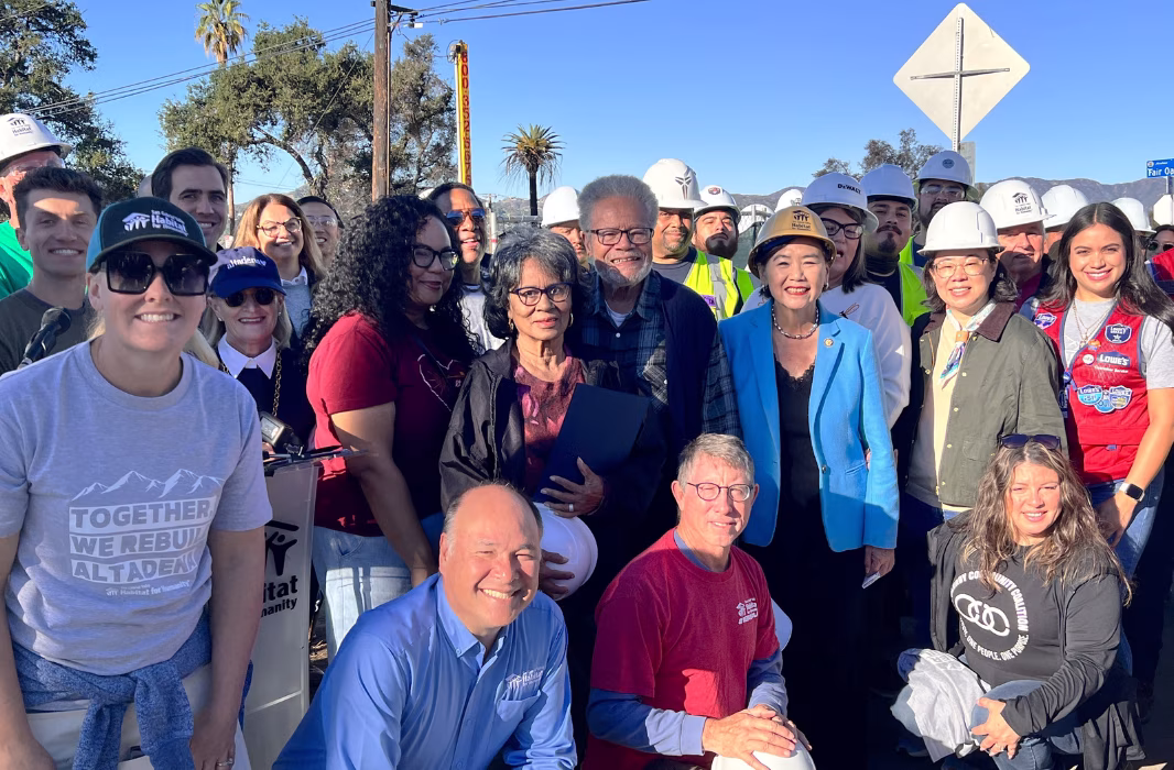 A large group of people stand outdoors under very blue skies, many are wearing construction hardhats.