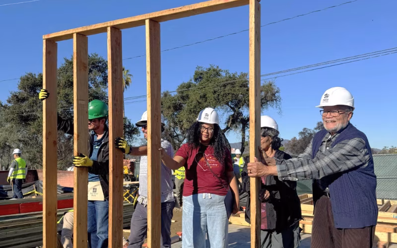 Five adults stand behind a framed wall at a construction site, helping raise it into position