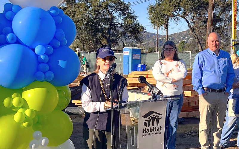 A woman in a baseball cap and sunglasses stands at a Habitat for Humanity-branded podium sharing remarks at a construction site. Two adults stand at the right of the photo listening to her remarks. 