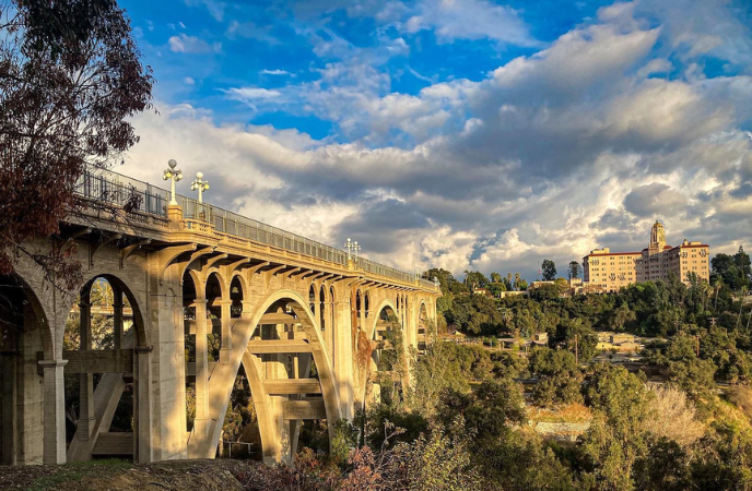 A sunset landscape photo of Pasadena's Colorado Street Bridge on the left, the historic Arroyo Vista Hotel on the right, and dramatic clouds in the sky