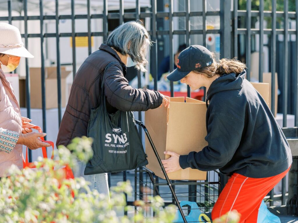 A young woman volunteer arranges a large box into a woman's wheeled cart at a food distribution