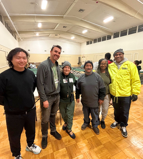 Six adults stand inside a gymnasium, several of them wearing outdoor clothing.