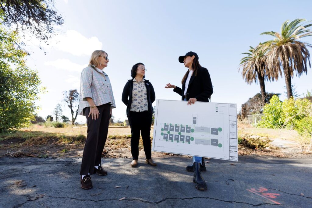 Three women stand on pavement outdoors discussing, one holds a large posted of architectural renderings. 