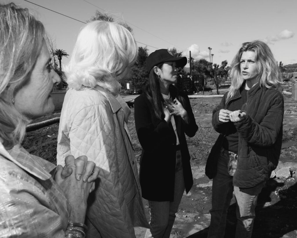 Four women stand together outdoors discussing, a dirt lot is in the background