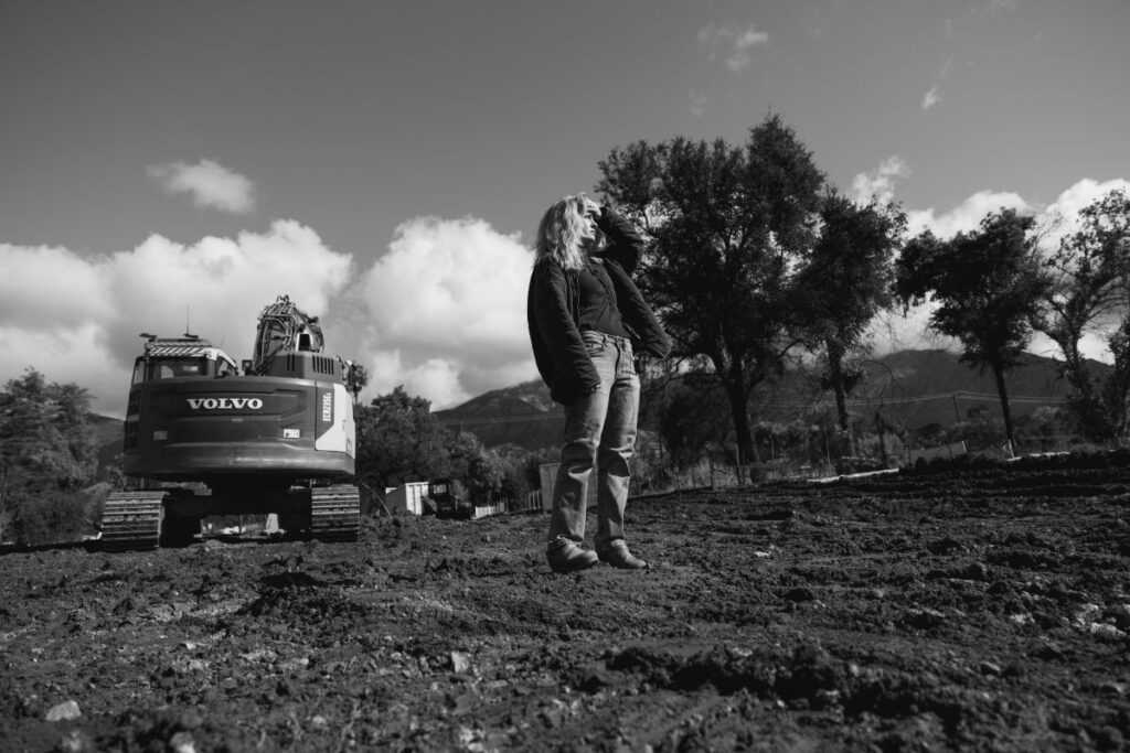 A black-and-white image, a woman standing on a dirt lot, shielding her eyes from the sun, an excavator behind her. 