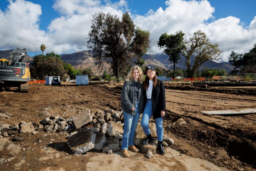 Two women stand together outside on a cleared, dirt lot with trees, clouds, and mountains behind them