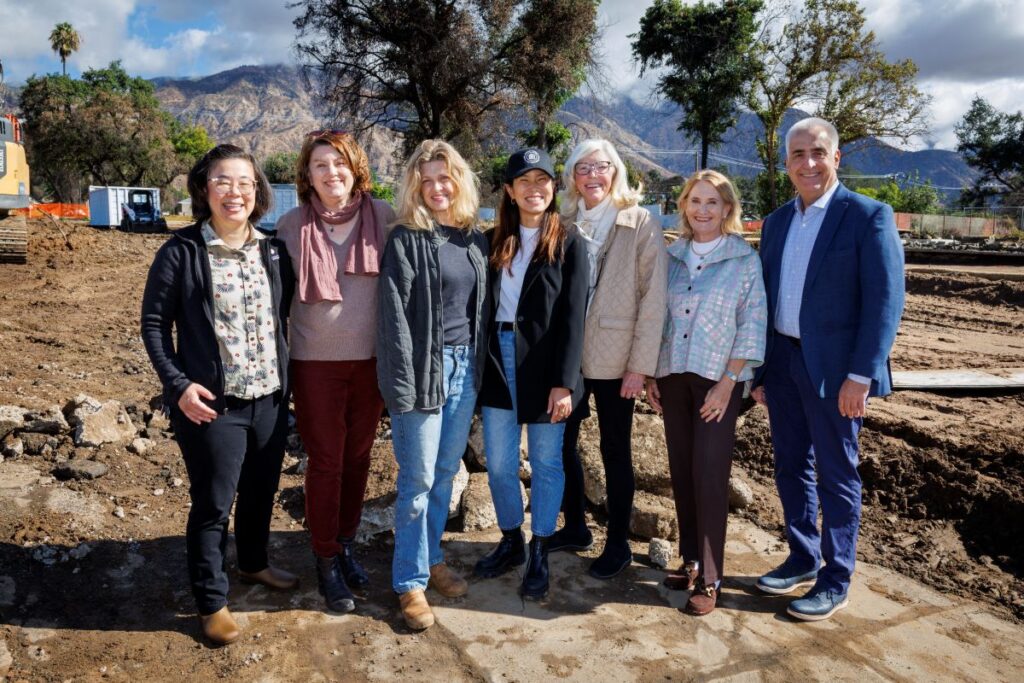 A group of 7 adults stand together, arm in arm, outdoors on a dirt lot with trees and mountains behind them