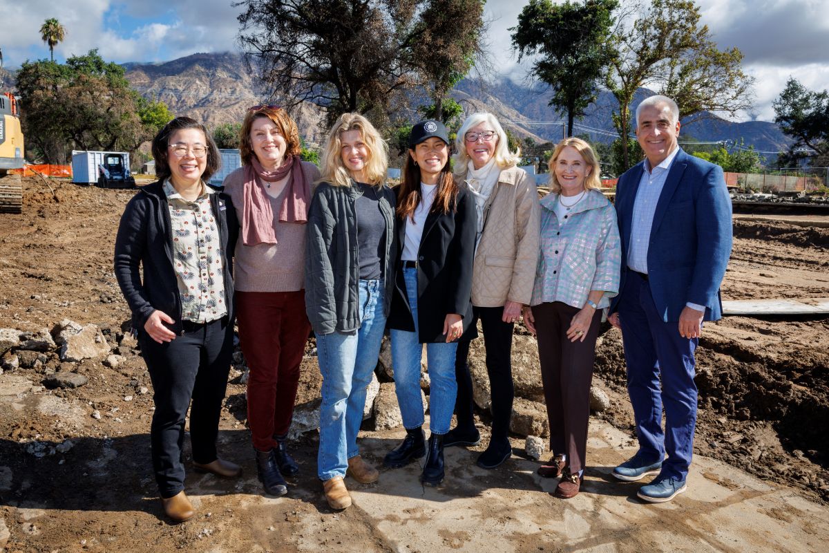 A group of 7 adults stand together, arm in arm, outdoors on a dirt lot with trees and mountains behind them