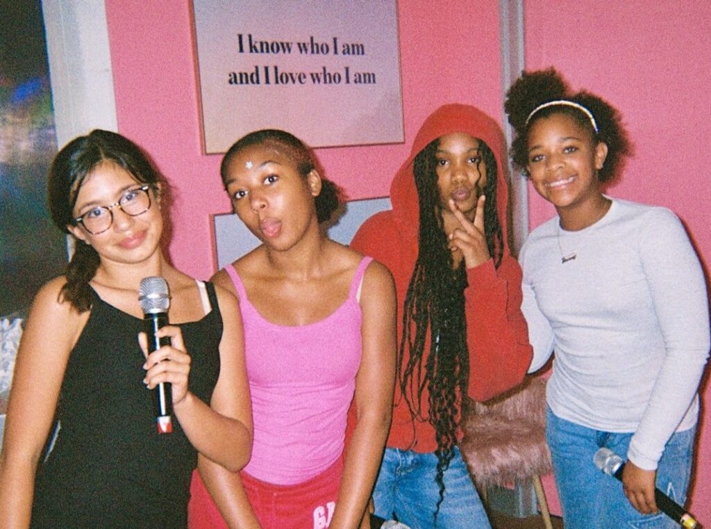 Four teen girls stand in front of a sign that reads I know who I am and I love who I am.