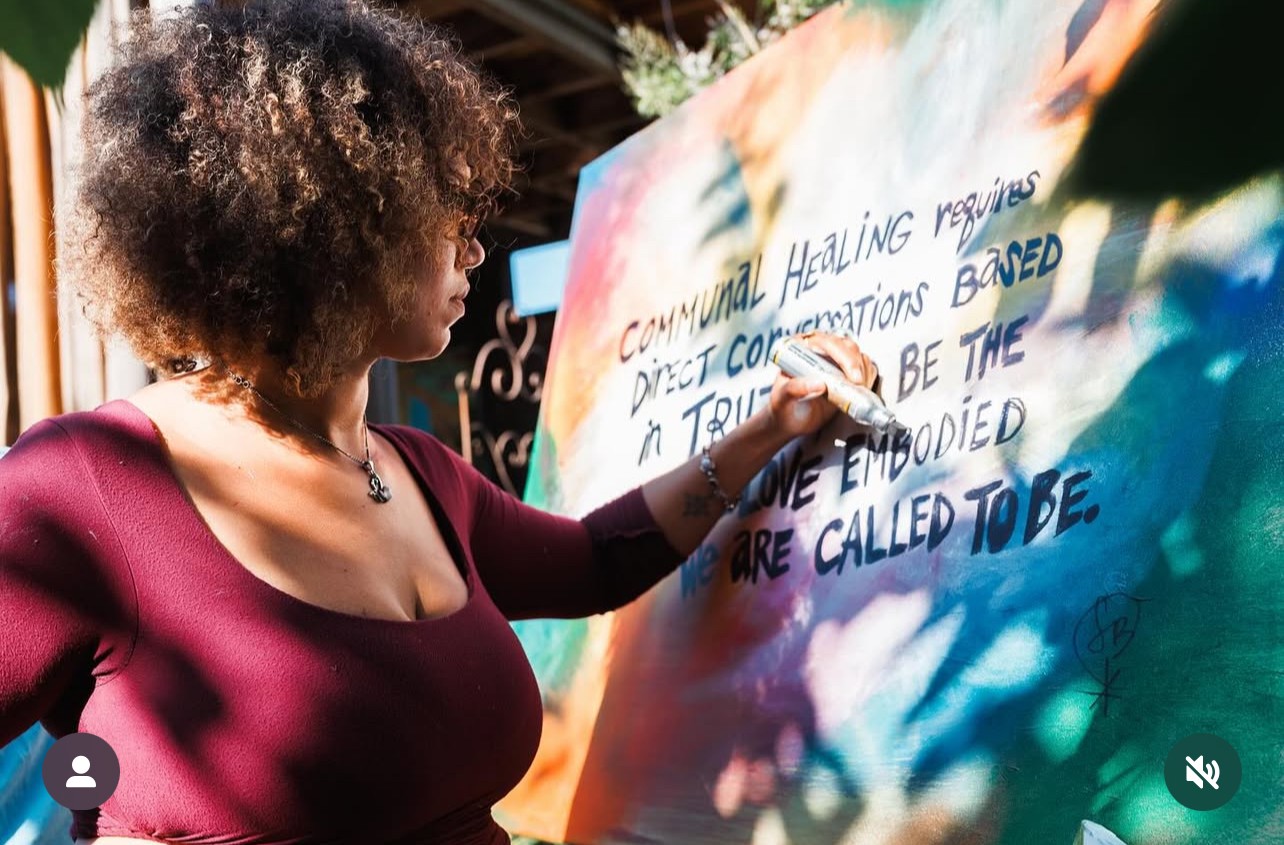A Black woman in a red shirt writes on a large colorful poster board