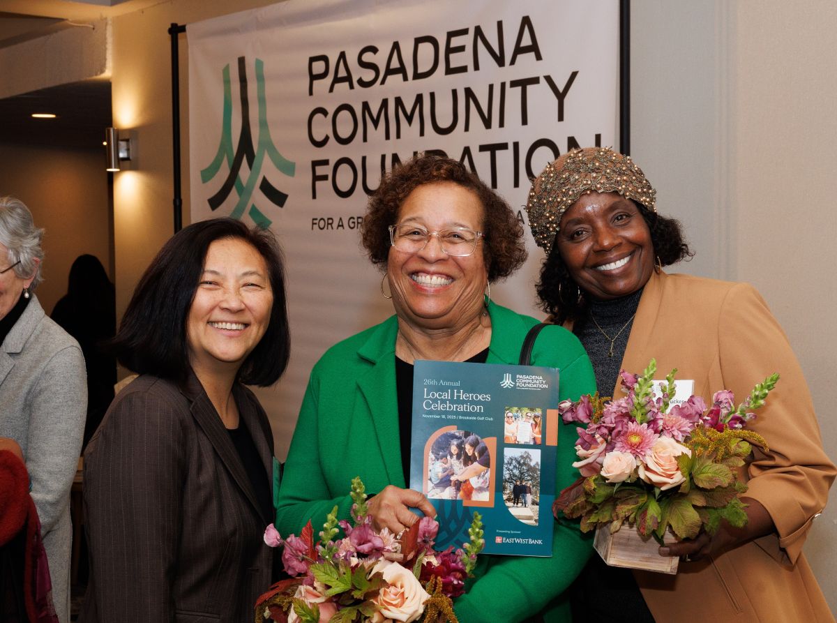 three African American women stand together holding floral arrangements and a program that reads Local Heroes Celebration. Behind them is a banner that says Pasadena Community Foundation
