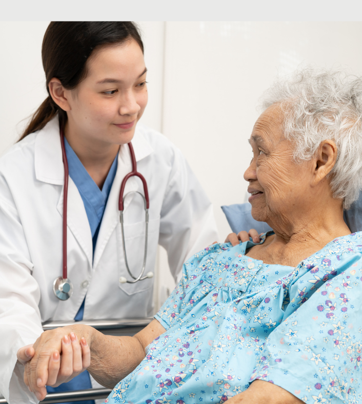 An Asian doctor stands closely to an elder Asian female patient dressed in an exam gown