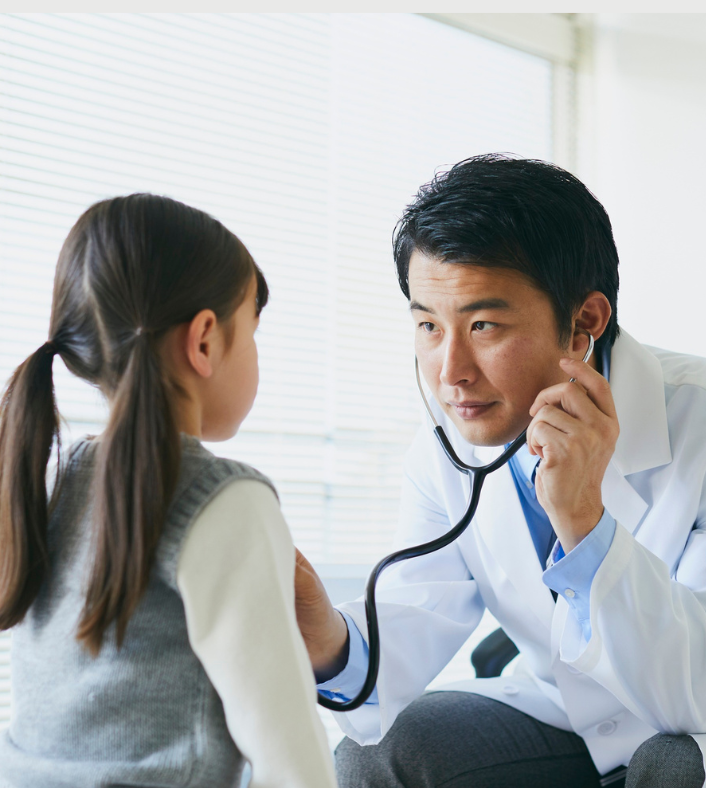 An Asian male doctor wearing a stethoscope leans forward to listen to a young girls heart