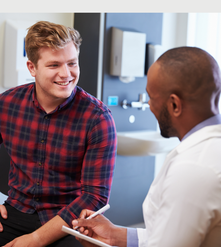 An anglo man in a red and black plaid shirt sits laughing and conversing with an African American doctor in a white coat