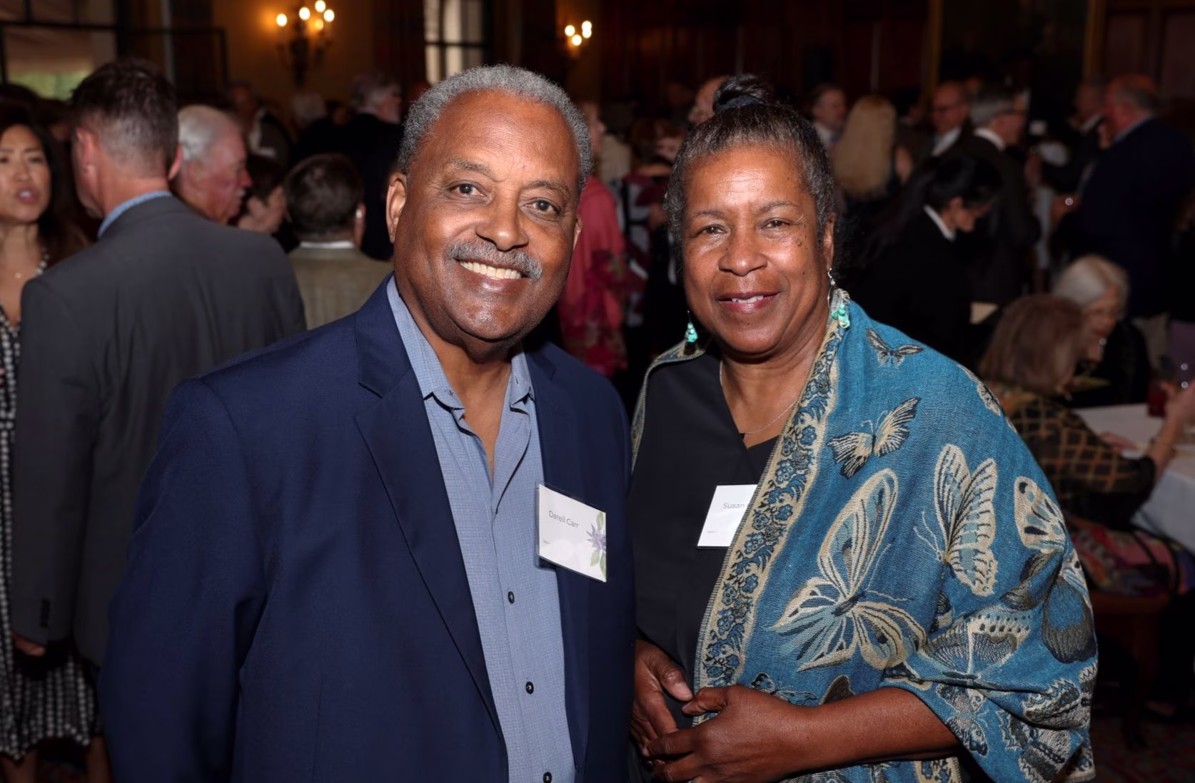 An African American middle age couple stand together at an event, people seen vageuly in the background