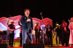 Taylor Goldsmith of Dawes stands at the forefront of a stage with his hand over his chest and near a microphone singing. Behind him are several more musical artists.
