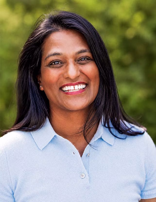 A woman is smiling in a blue button down shirt with a green leaf background.
