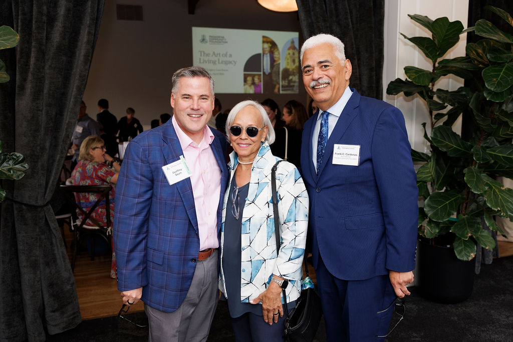 3 adults standing together. Two men in blue blazers and a woman in a floral blazer with sun glasses on