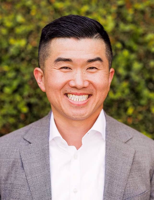 A man in a grey suit is smiling. Green leaf background.