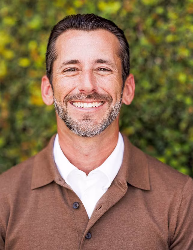 A man in a rust color button down is smiling. Green leaf background.
