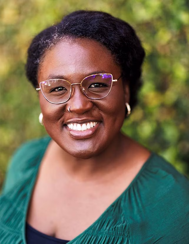 A woman in a green shirt with her hair in a bun is smiling. She is wearing gold earrings. Green leaf background.