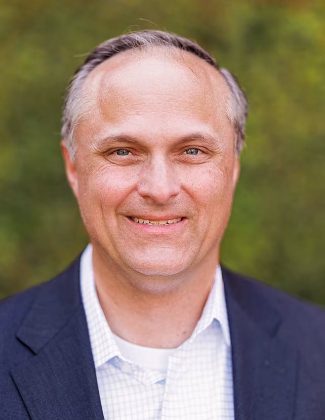 A man in a blue suit and white Botton up shirt is smiling. Green leaf background.