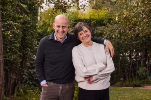 A man and a woman are facing the camera and smiling in nature. They are happy and hugging from the side.